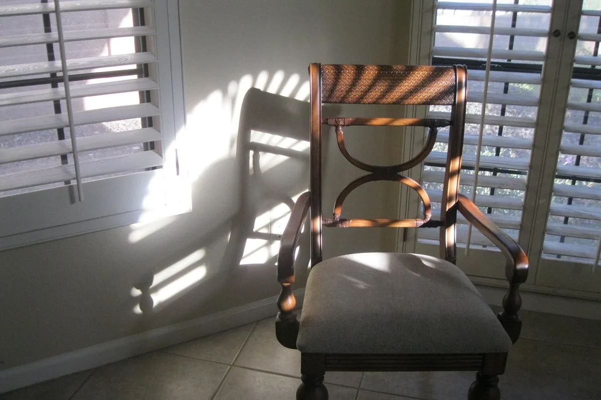 Rayos de sol entrando por unas contraventanas sobre una silla de madera y un suelo de baldosas