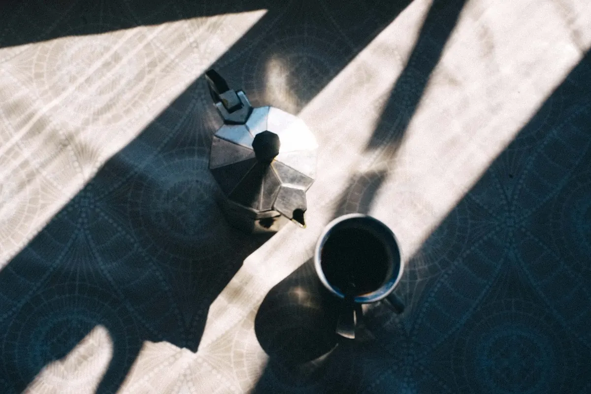 Cafetera italiana de aluminio y una taza de café negro sobre un mantel con dibujos, bañados por la luz de una ventana