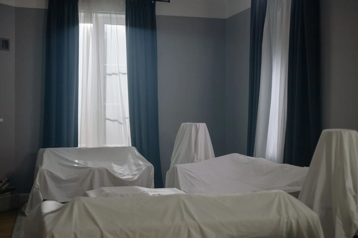 Salón de una vivienda cerrada con los sofás y las mesas cubiertos con sábanas blancas, junto a una ventana con cortinas azules