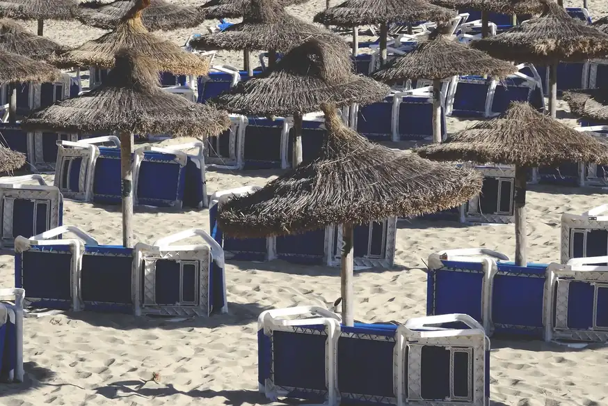 Sombrillas de paja y tumbonas azules apiladas sobre la arena de una playa sin gente