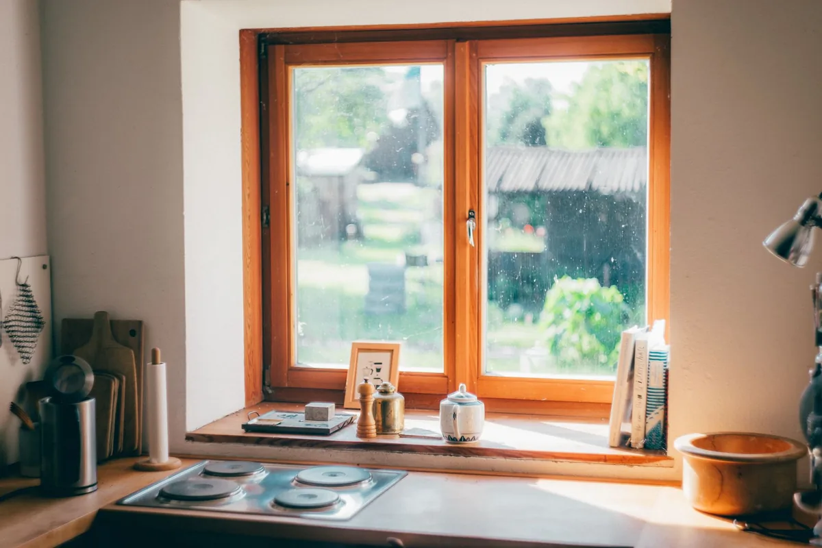 Cocina con una ventana de madera por la que entra el sol, con una placa eléctrica, tablas de cortar y una tetera en el alféizar