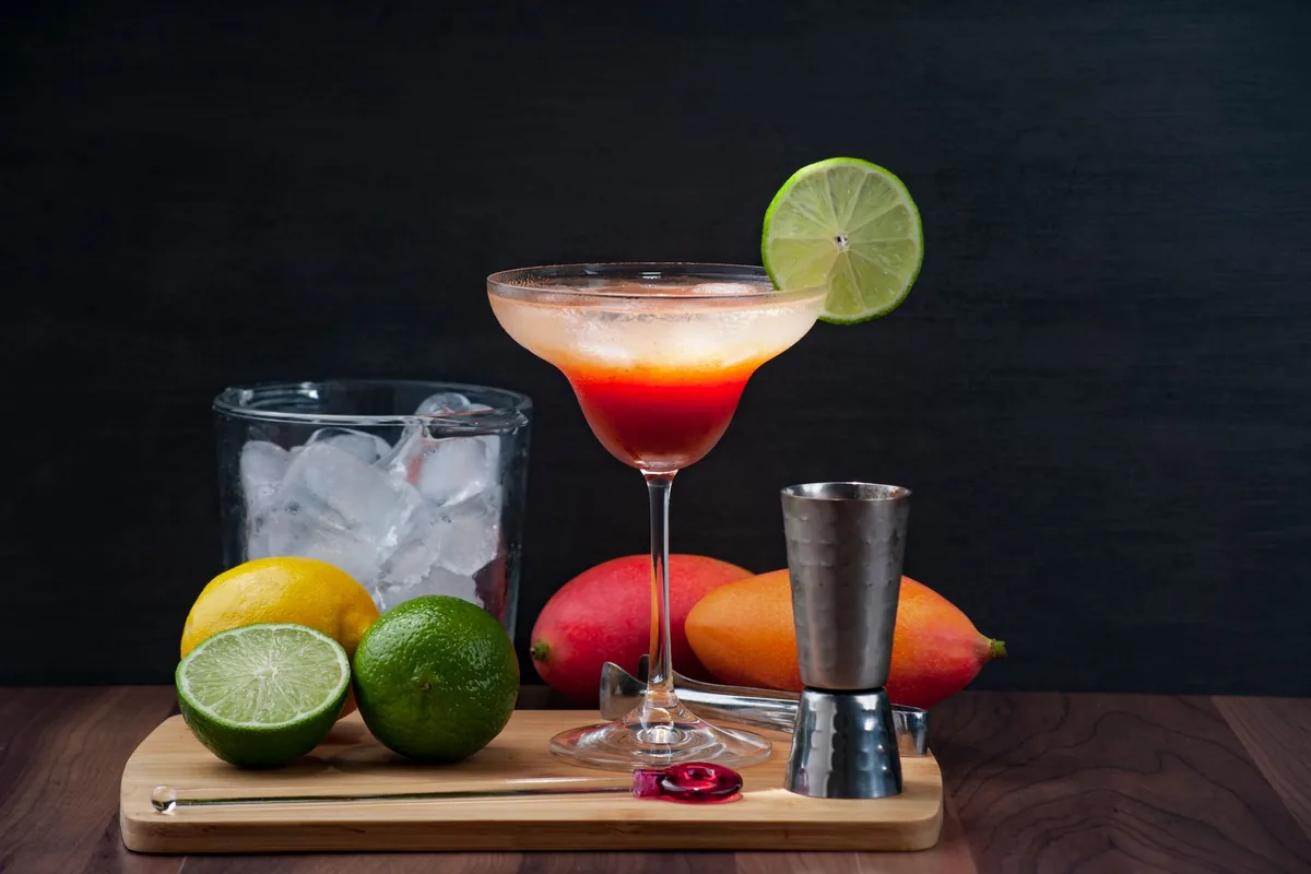 Cubo de cristal con hielo, un limón, dos limas y dos mangos junto a una copa con una bebida roja y un medidor de acero, sobre una tabla de madera