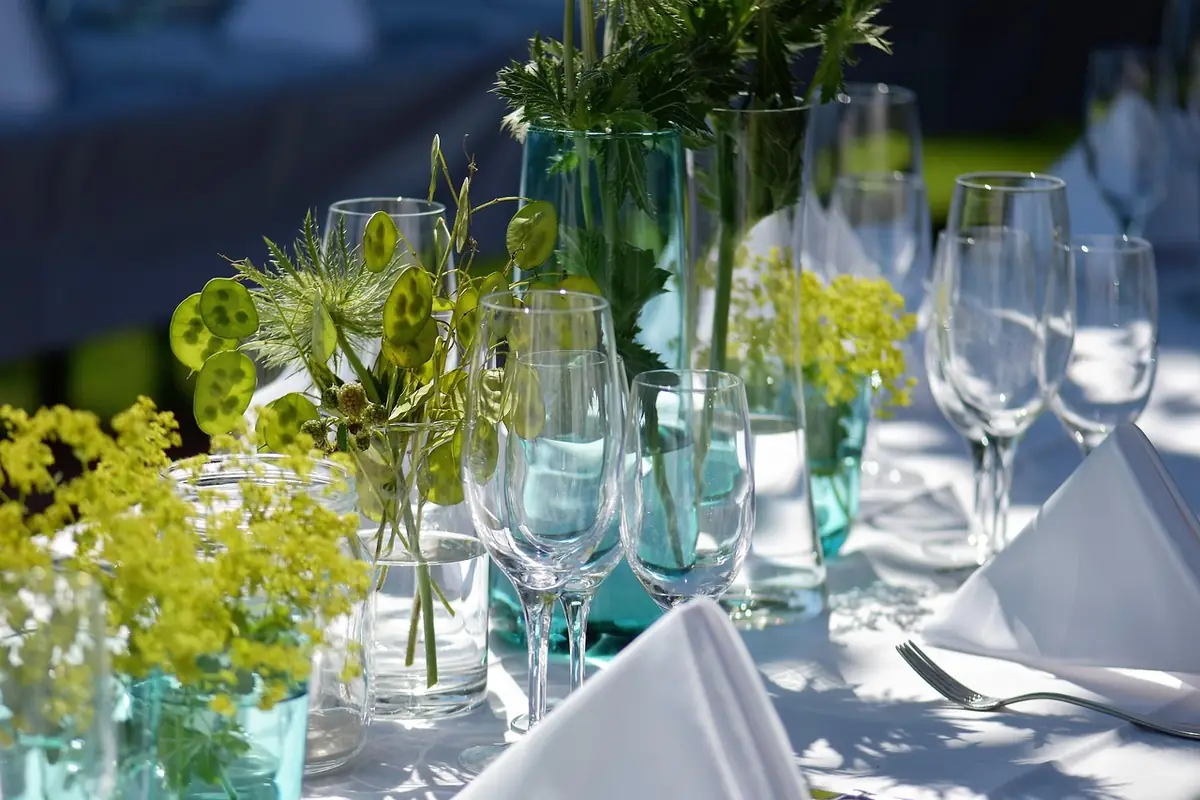 Mesa puesta al aire libre con copas de cristal, jarrones con flores amarillas y verdes y servilletas blancas, a pleno sol