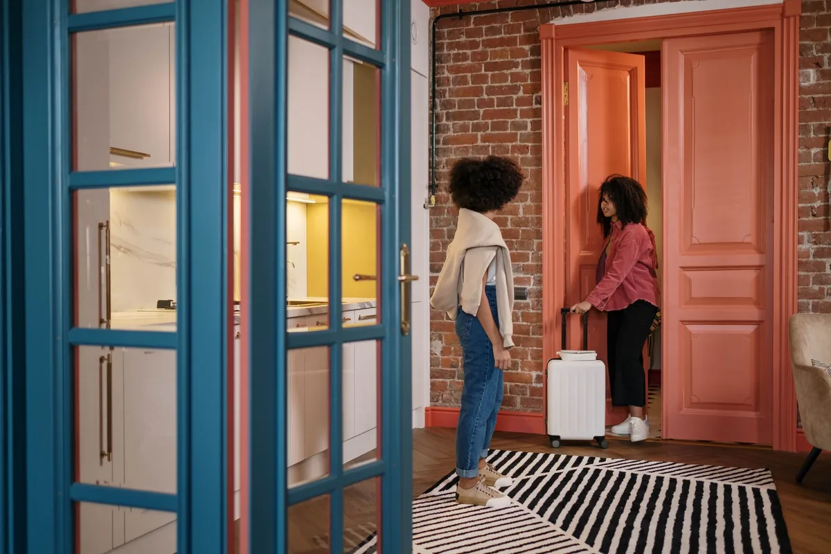 Dos personas junto a la puerta de un apartamento, una con una maleta blanca, y la cocina al fondo tras una puerta acristalada azul