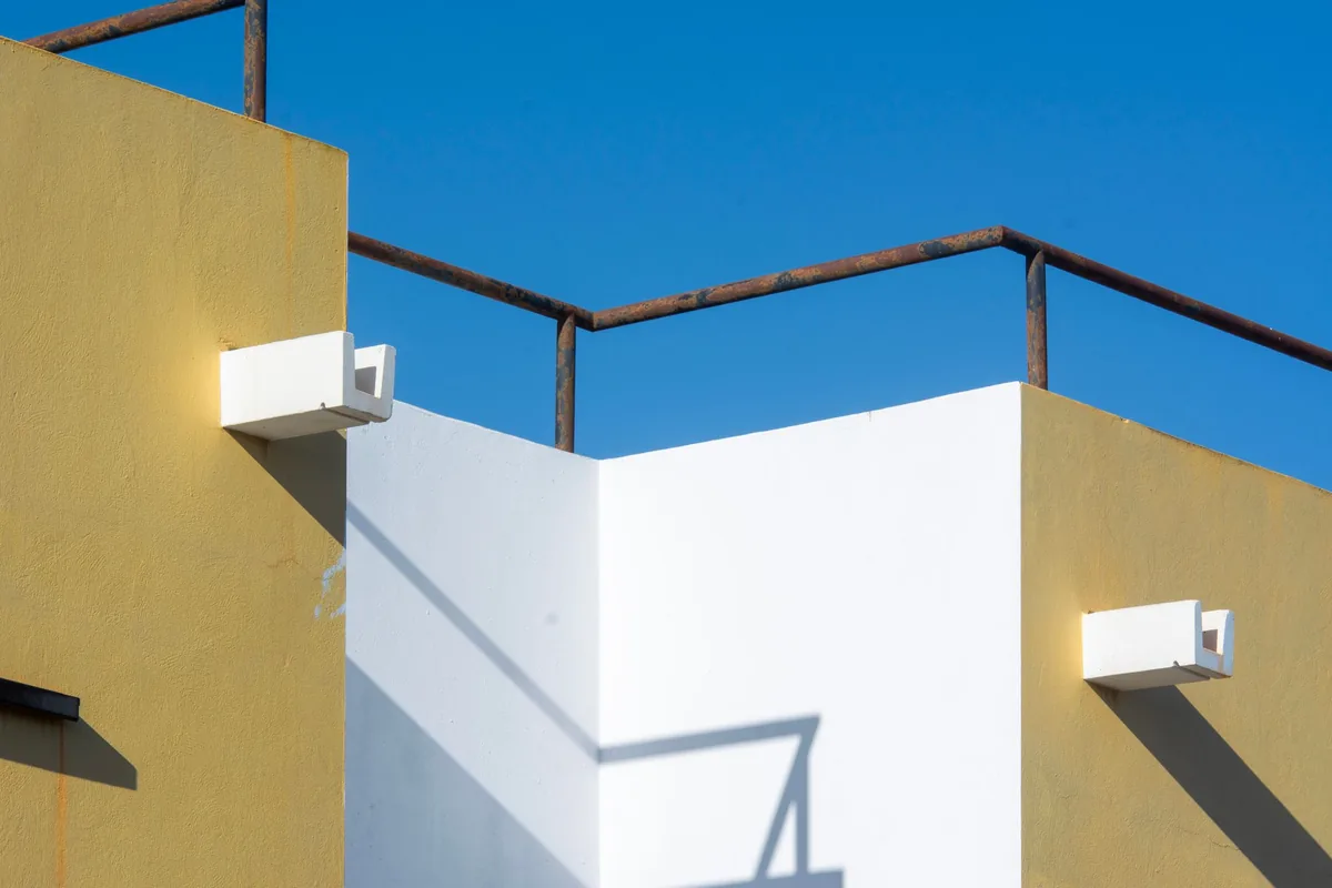 Esquina de una azotea con muros amarillos y blancos y una barandilla oxidada bajo un sol fuerte y un cielo azul