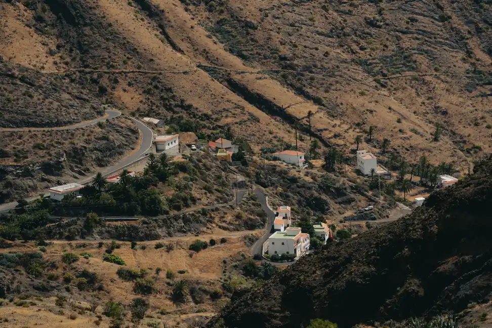 Casas blancas de tejados naranjas entre palmeras en una ladera seca y dorada por el sol, con una carretera que serpentea