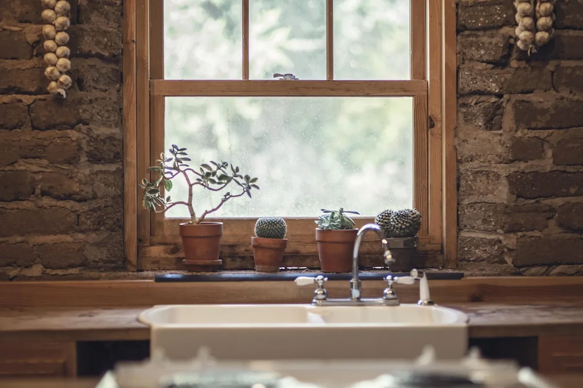 Ventana de madera sobre un fregadero blanco, con cuatro macetas de cactus y suculentas en el alféizar y una pared de ladrillo rústico