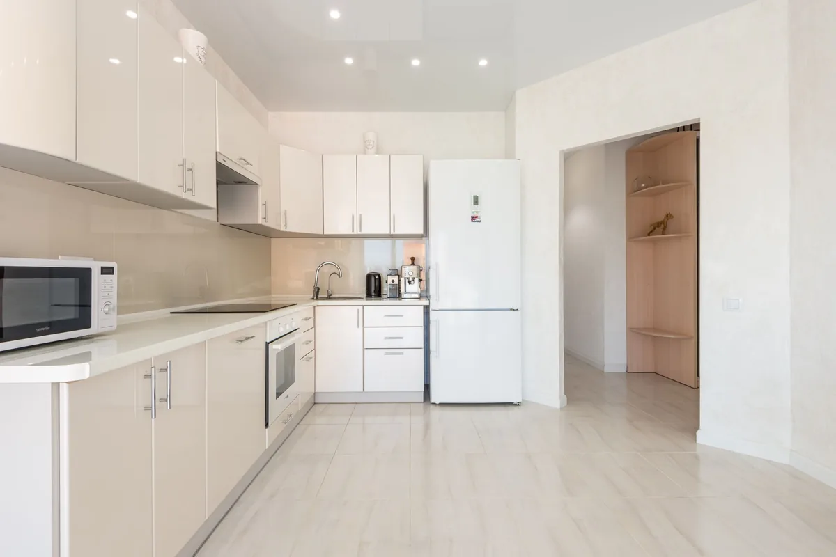 Cocina blanca luminosa con encimera, horno, microondas y un frigorífico blanco de dos puertas junto al paso al pasillo