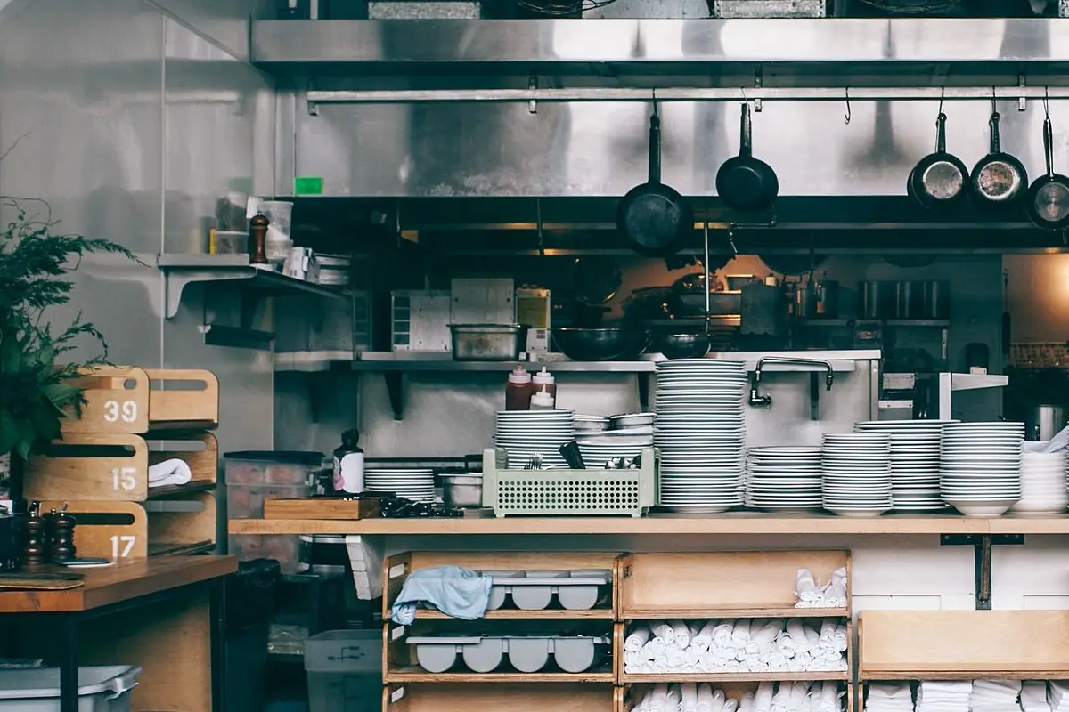 Cocina de un restaurante con campana de acero, sartenes colgadas y estantes con pilas de platos y servilletas