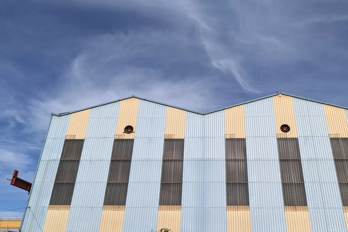 Fachada de una nave industrial de chapa azul clara y amarilla con ventanales oscuros y dos ventiladores bajo un cielo de nubes finas
