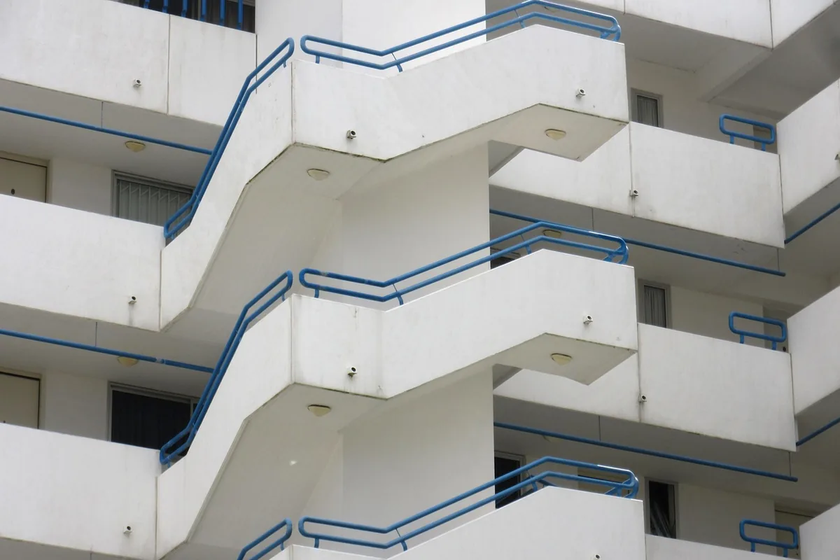 Fachada blanca de un edificio de apartamentos con escaleras exteriores y barandillas azules, vista desde abajo
