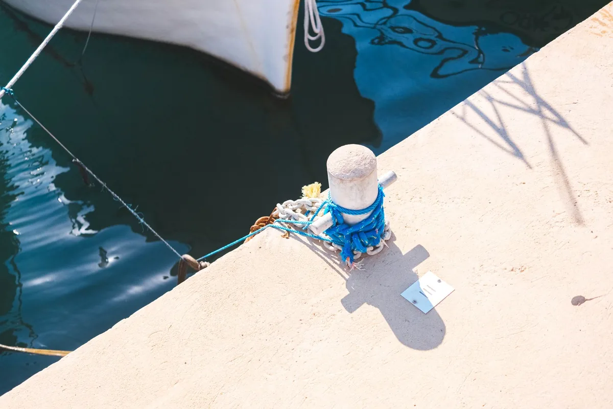 Noray de hormigón en el borde de un muelle con una cuerda azul enrollada, junto al agua y la proa de una barca blanca