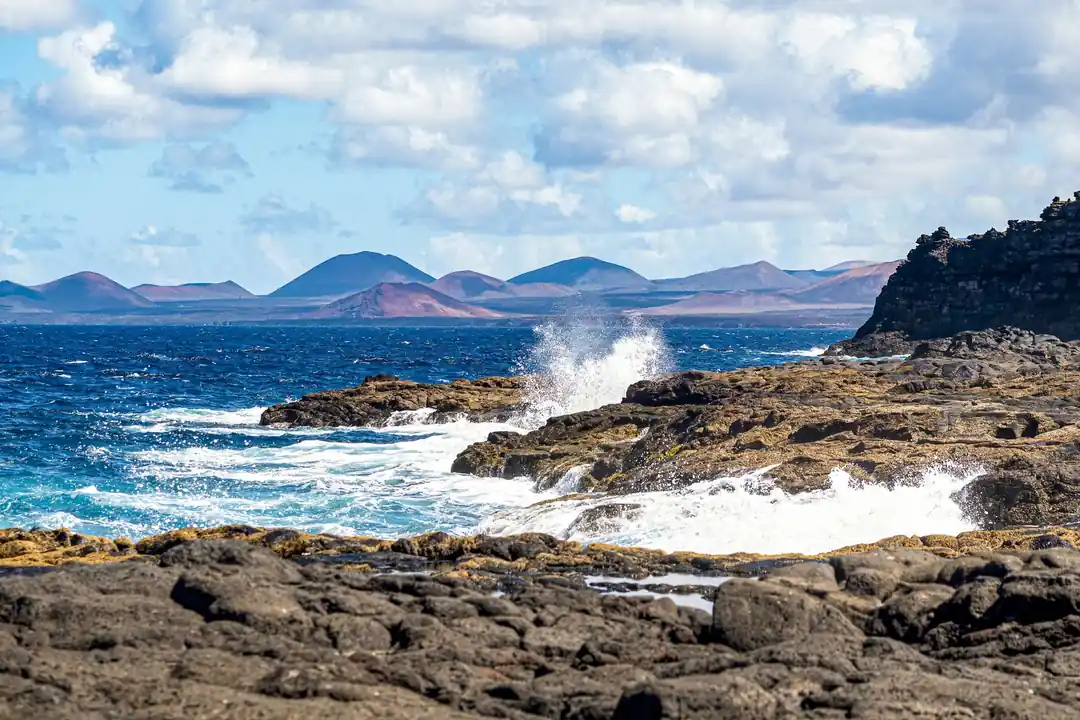 Olas rompiendo contra una costa de rocas volcánicas oscuras, con montañas rojizas al fondo bajo un cielo de nubes