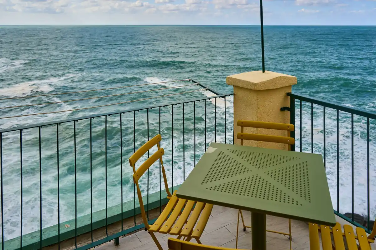 Balcón con una mesa verde y sillas plegables amarillas junto a una barandilla, con el mar agitado y espuma blanca al fondo