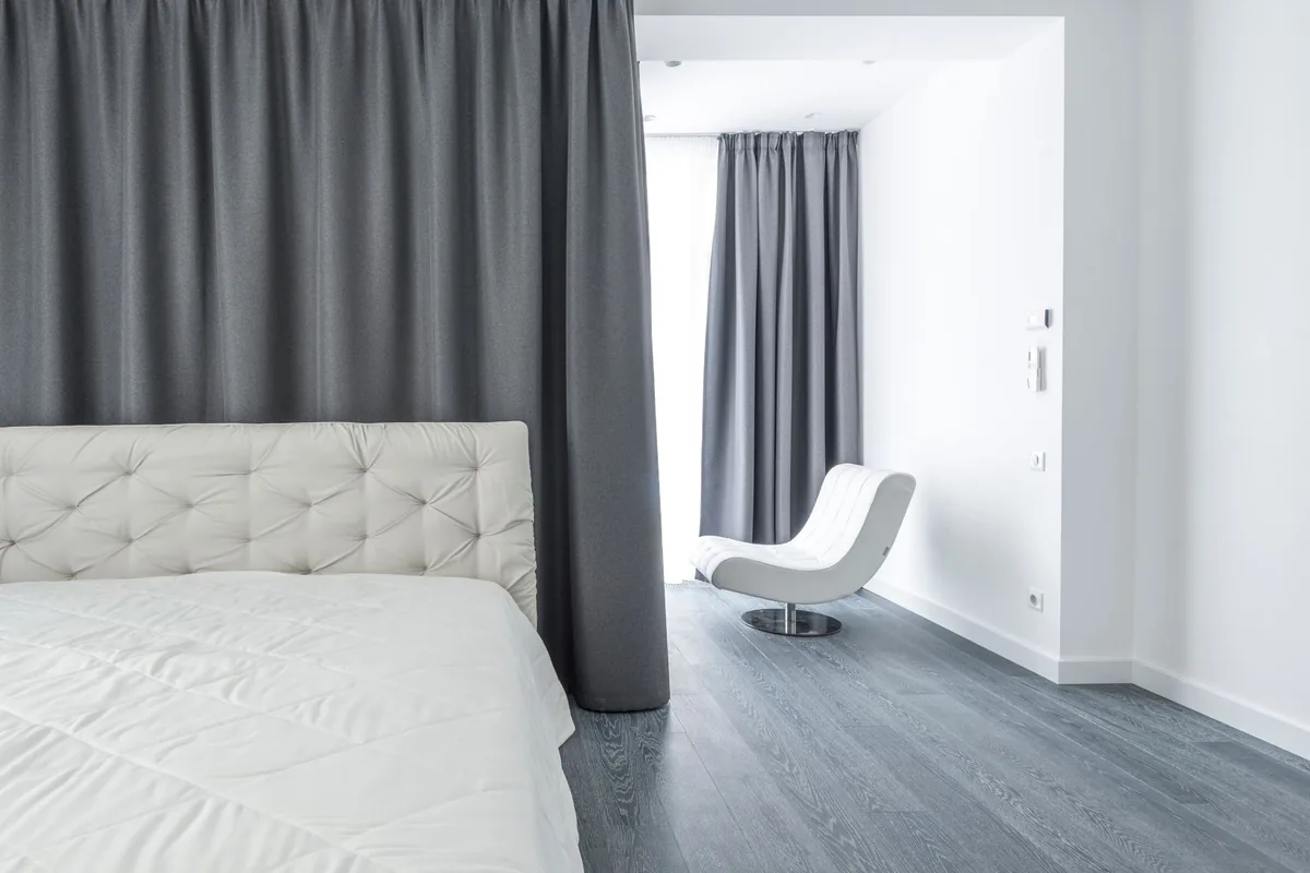 Dormitorio blanco con una cama de cabecero acolchado, cortinas grises hasta el techo y un sillón blanco al fondo