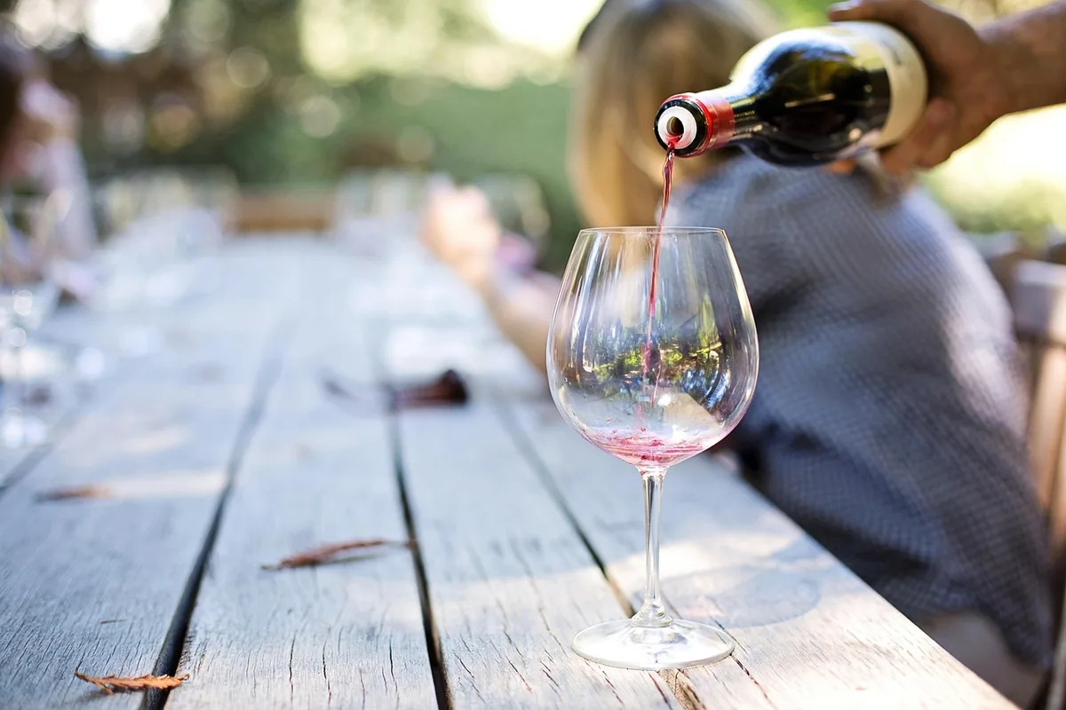 Una mano sirve vino tinto en una copa sobre una mesa larga de madera al aire libre