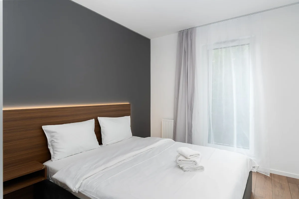 Cama hecha con sábanas blancas y tres toallas dobladas, junto a una ventana con cortinas grises y visillo, en un dormitorio de alojamiento