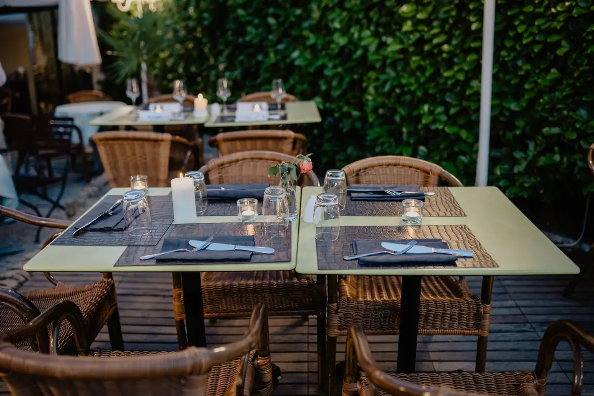 Mesas de una terraza de restaurante con cubiertos, vasos y velas puestos, sillas de mimbre y un seto verde al fondo, al caer la tarde