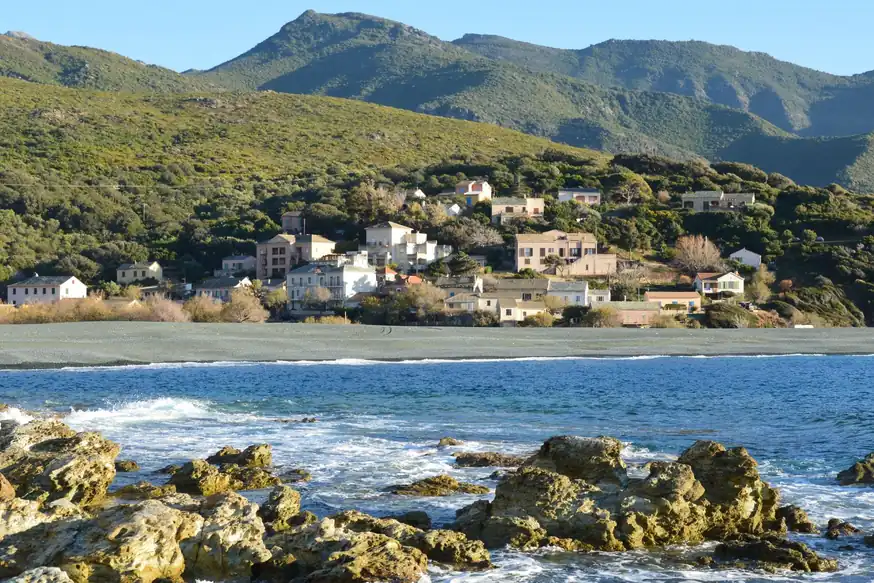 Pueblo de casas claras al pie de montañas verdes, con una playa de guijarros oscuros y rocas con el mar en primer término